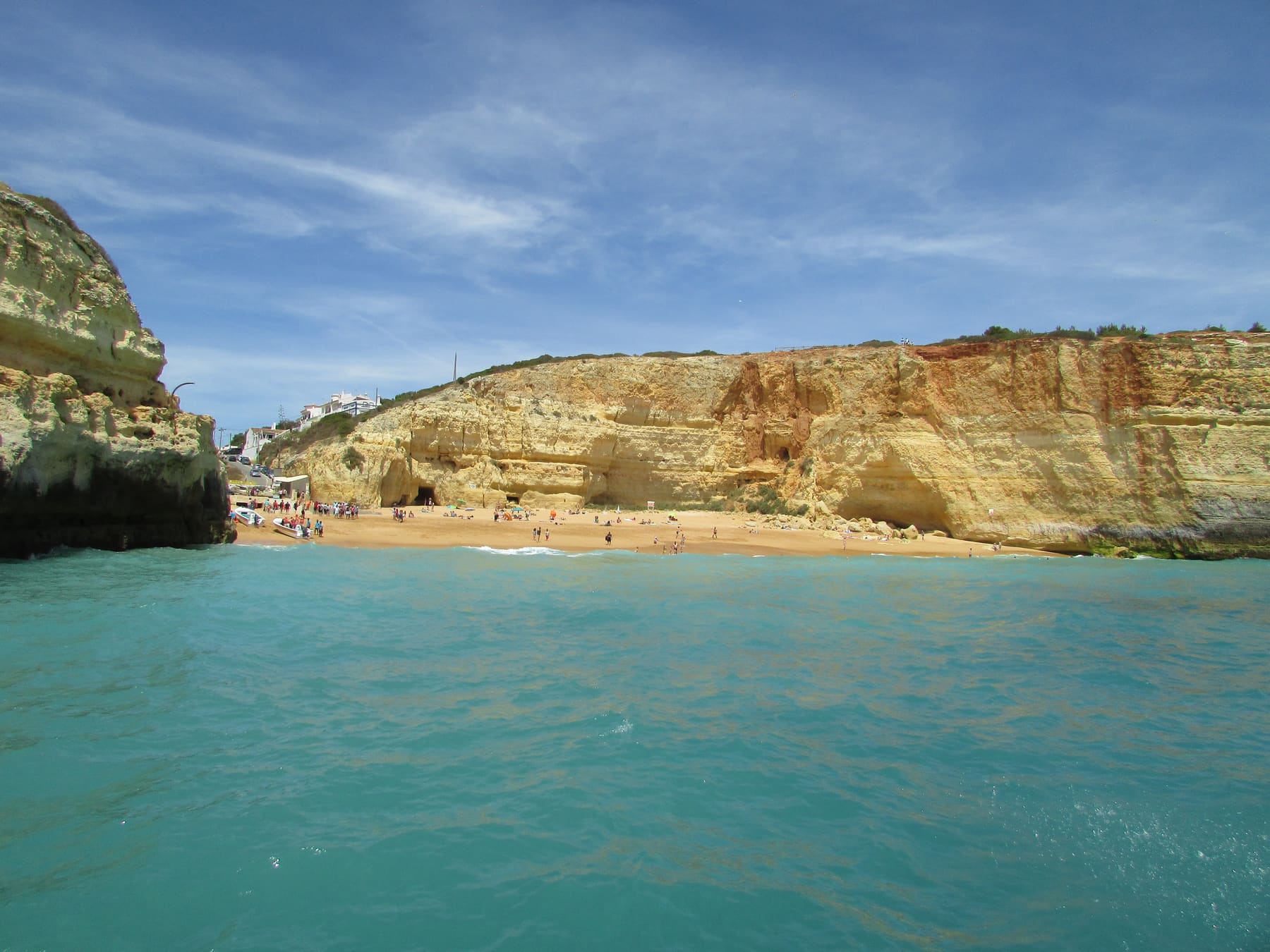 La Praia de Benagil vista desde el mar: una pequeña playa de arena entre acantilados ocres, con casas del pueblo encima.